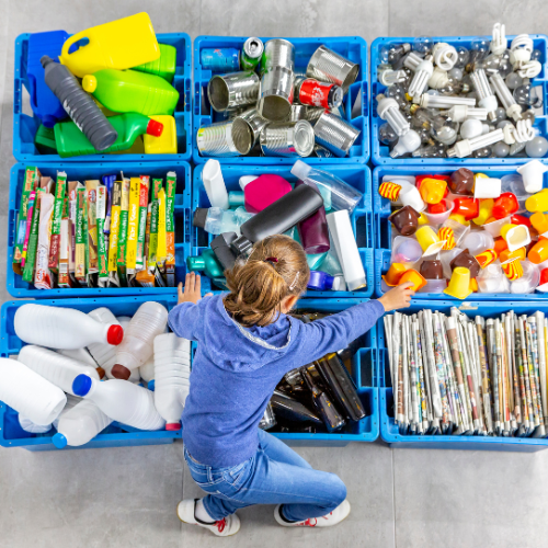 Recycling education: Child sorting plastics, metal cans, paper, and cartons into colour-coded recycling bins indoors.